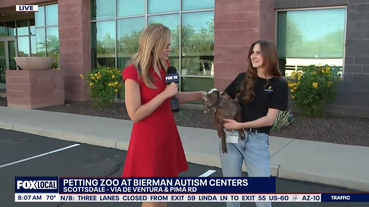 A Fox 10 Arizona news reporter in a red dress interviews a Bierman Autism Centers team member holding a small brown goat outside the Scottsdale center. Yellow flowers and a red brick building are visible in the background.