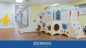 Indoor therapy playroom at Bierman Autism Centers featuring a wooden climbing structure with a slide, balance wall, and blue penguin footprints on a clean, wood-style floor.