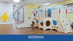 Indoor therapy playroom at Bierman Autism Centers featuring a wooden climbing structure with a slide, balance wall, and blue penguin footprints on a clean, wood-style floor.