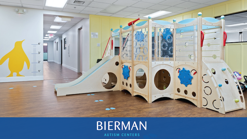 Indoor therapy playroom at Bierman Autism Centers featuring a wooden climbing structure with a slide, balance wall, and blue penguin footprints on a clean, wood-style floor.