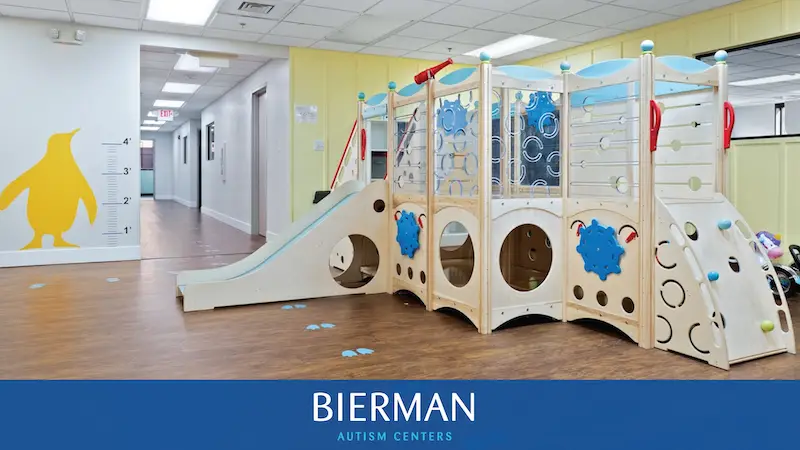 Indoor therapy playroom at Bierman Autism Centers featuring a wooden climbing structure with a slide, balance wall, and blue penguin footprints on a clean, wood-style floor.
