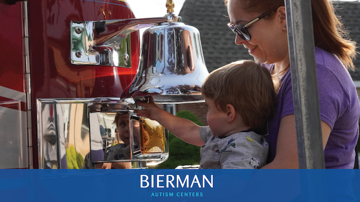 Mother supports toddler ringing a quiet fire-engine bell at Bierman Autism Centers’ sensory-friendly Touch-a-Truck event.
