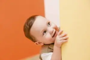 Smiling toddler peeks out from behind a wall during playful ABA therapy activity at home.