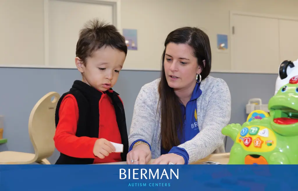 A Bierman Autism Centers clinician sits with a young boy at a therapy table, pointing to a card of colored shapes during an early learning activity.