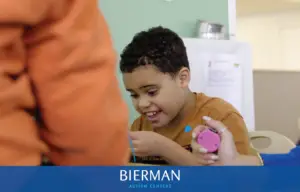 A smiling child works on a hands-on activity at a table while a therapist holds a timer during an ABA therapy session.