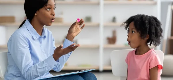 Attractive young african american woman speech therapist working with little black girl, sitting at table with colorful letters, learning pronunciation, clinic interior. Speech disorders in children